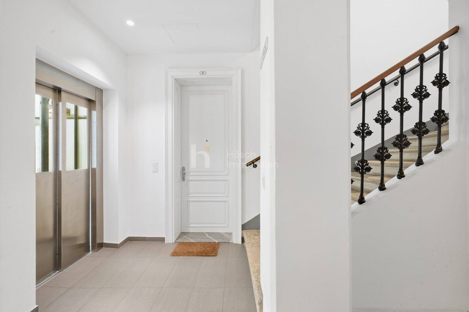 Eingangsbereich mit Aufzug und einer eleganten Treppe mit schmiedeeisernem Geländer.