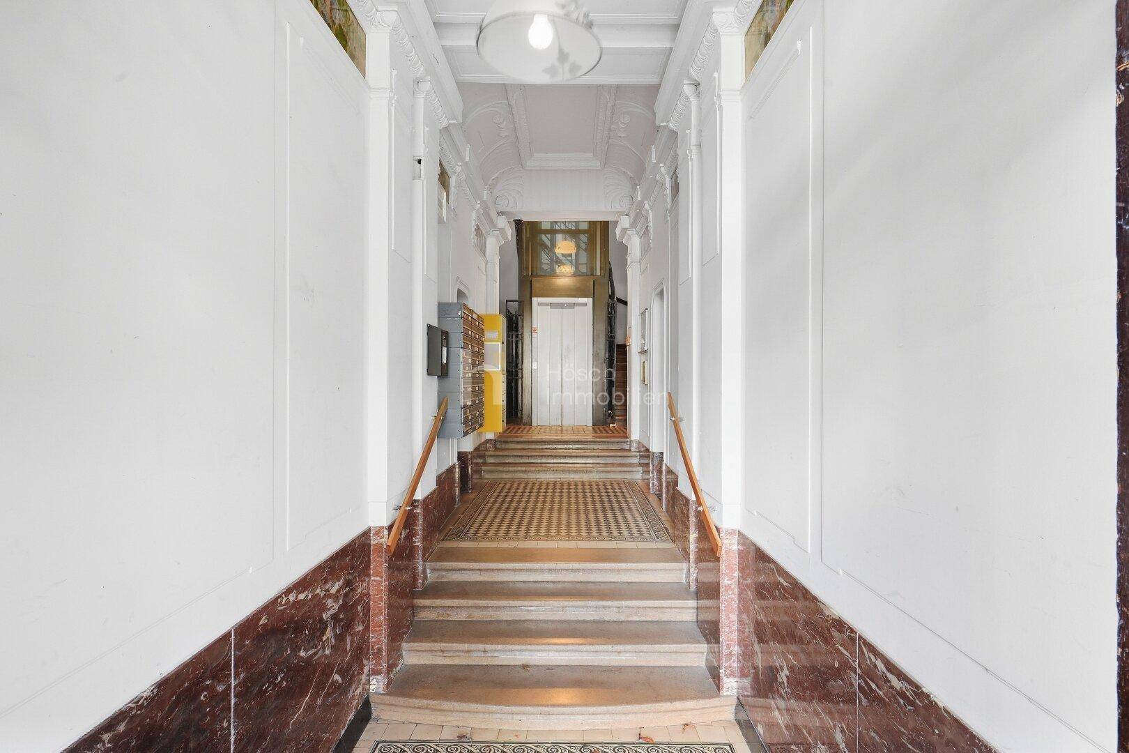 Klassischer Eingangsbereich mit breiter Treppe, Marmorwänden und einem historischen Aufzug im Hintergrund.