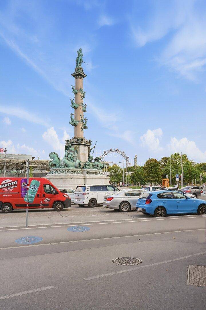 Belebtes Stadtbild mit einem historischen Denkmal, fahrenden Autos und dem Riesenrad im Hintergrund.