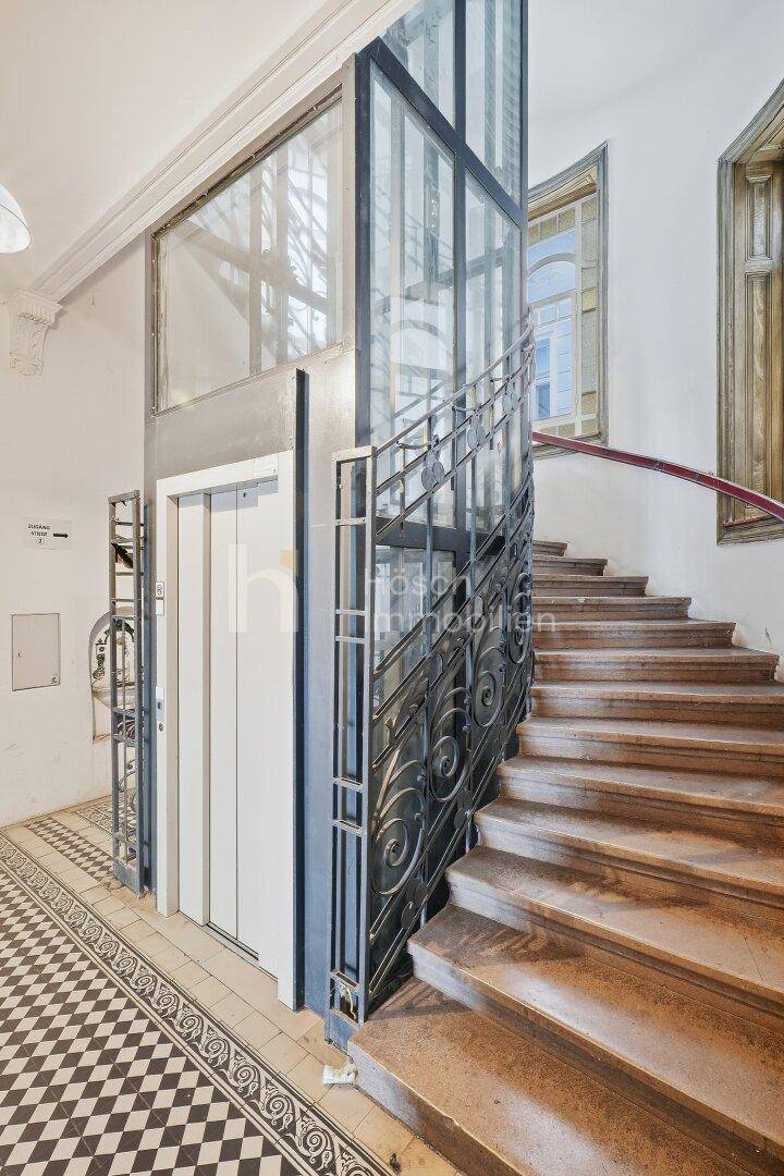 Elegantes Treppenhaus mit Glasaufzug und schmiedeeisernem Geländer, das den historischen Charme bewahrt.
