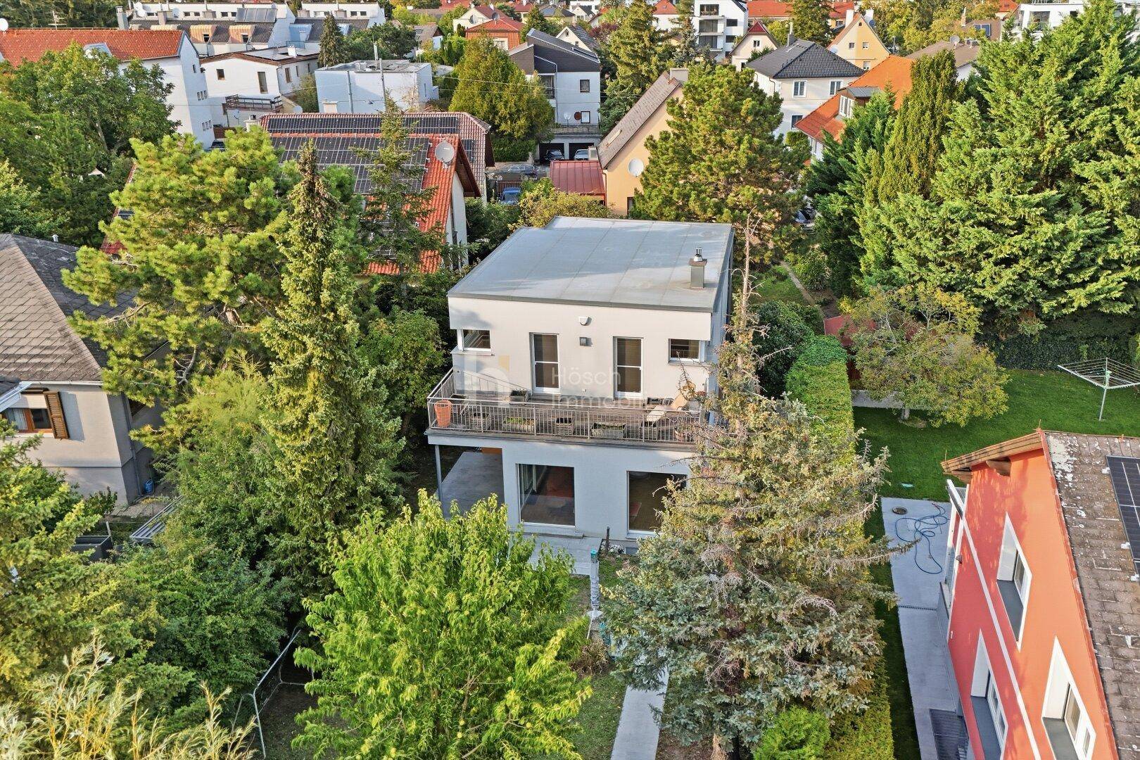 Luftaufnahme eines modernen Hauses mit Flachdach, umgeben von viel Grün und Nachbargebäuden.