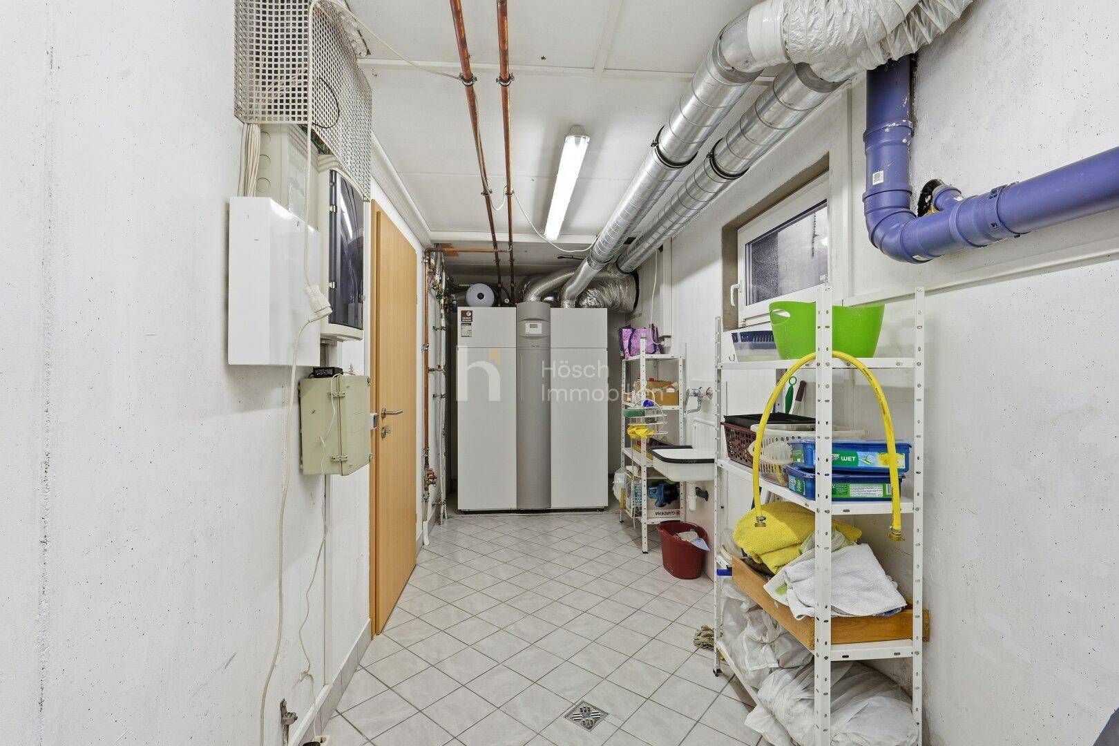 Technikraum im Keller mit Wärmepumpe, Rohrleitungen und zusätzlichen Regalen.