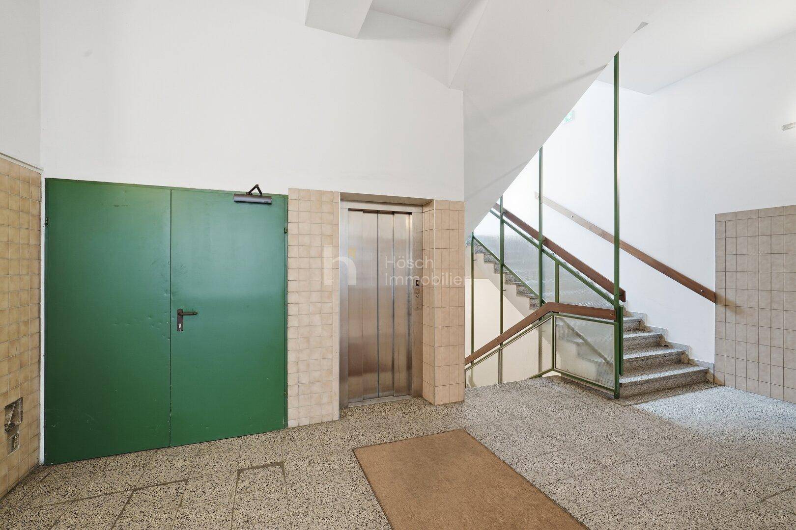 Treppenhaus mit Terrazzoboden, Geländer und einem modernen Aufzug im Eingangsbereich.