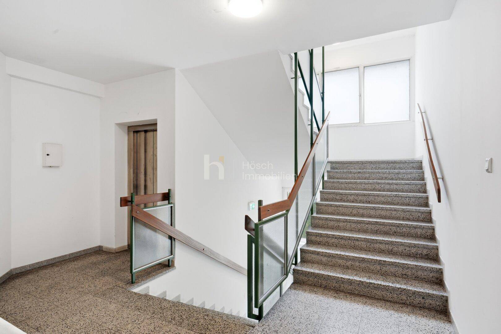 Treppenhaus mit Terrazzoboden, Geländer und einem sichtbaren Aufzug im Hintergrund.