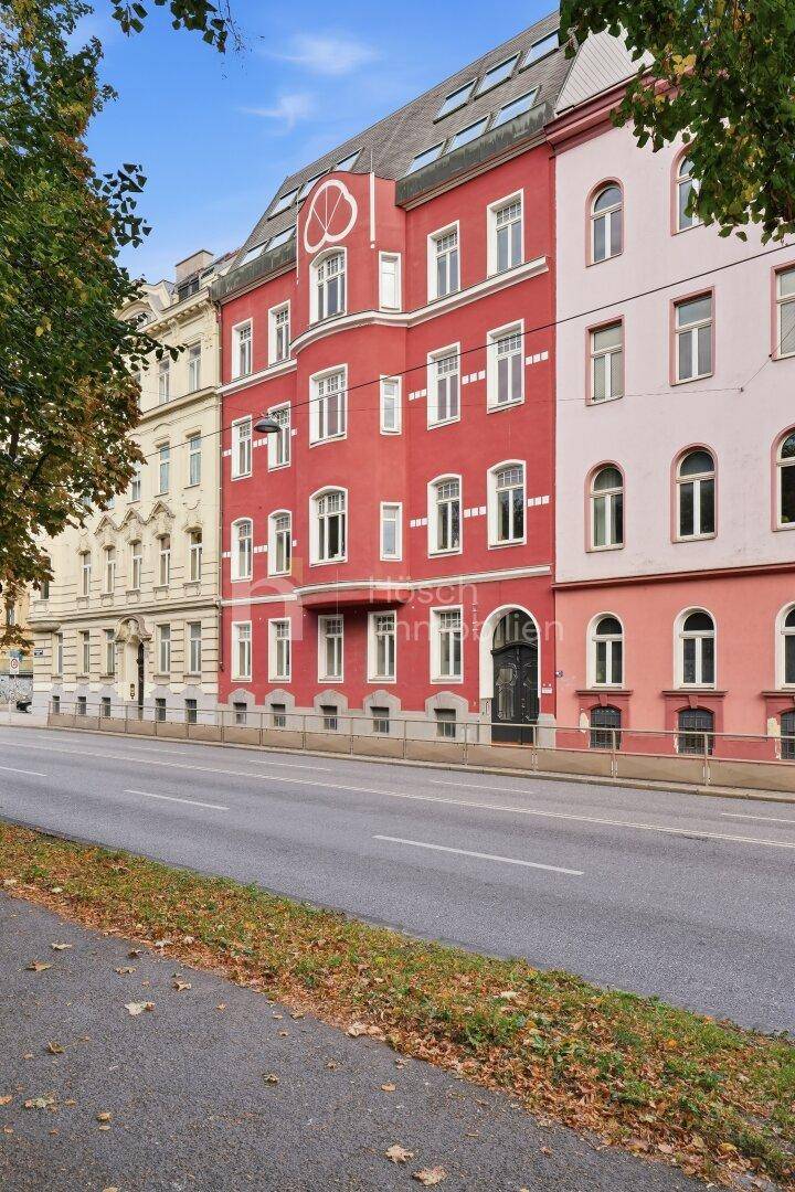 Rotes Mehrfamilienhaus mit weißen Fenstern an einer belebten Straße, umgeben von weiteren Gebäuden.