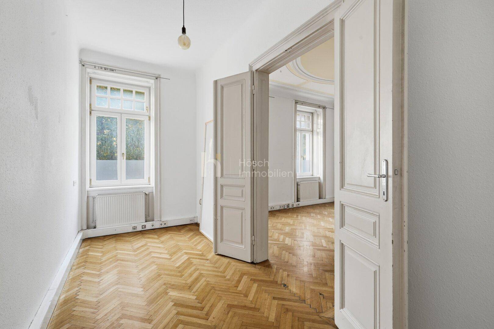 Heller Flur mit Fischgrätparkett und hohen Türen, der zu einem weiteren Zimmer führt.