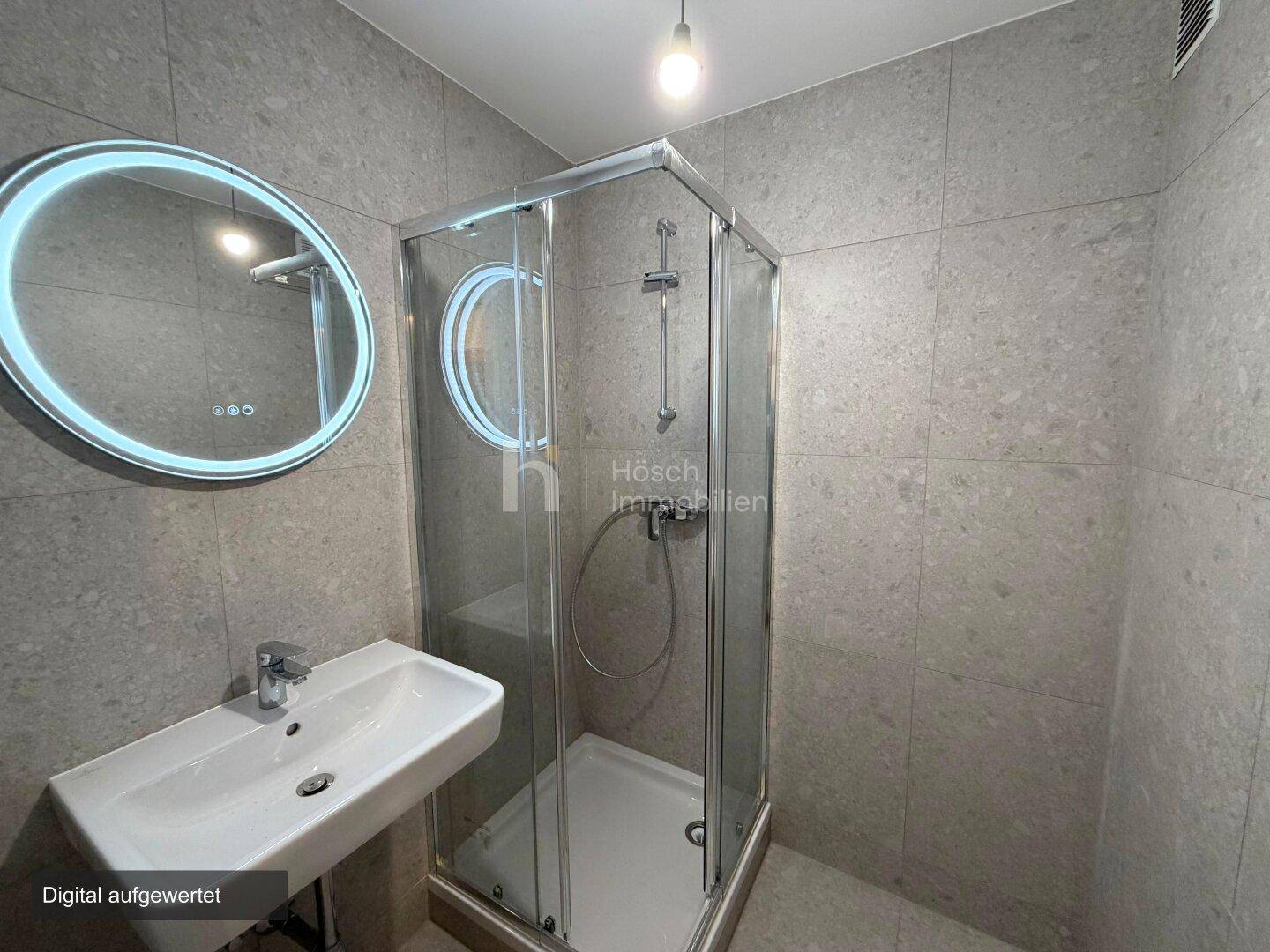 Modernes Badezimmer mit grauen Fliesen, runder Spiegelleuchte und einer Duschkabine.