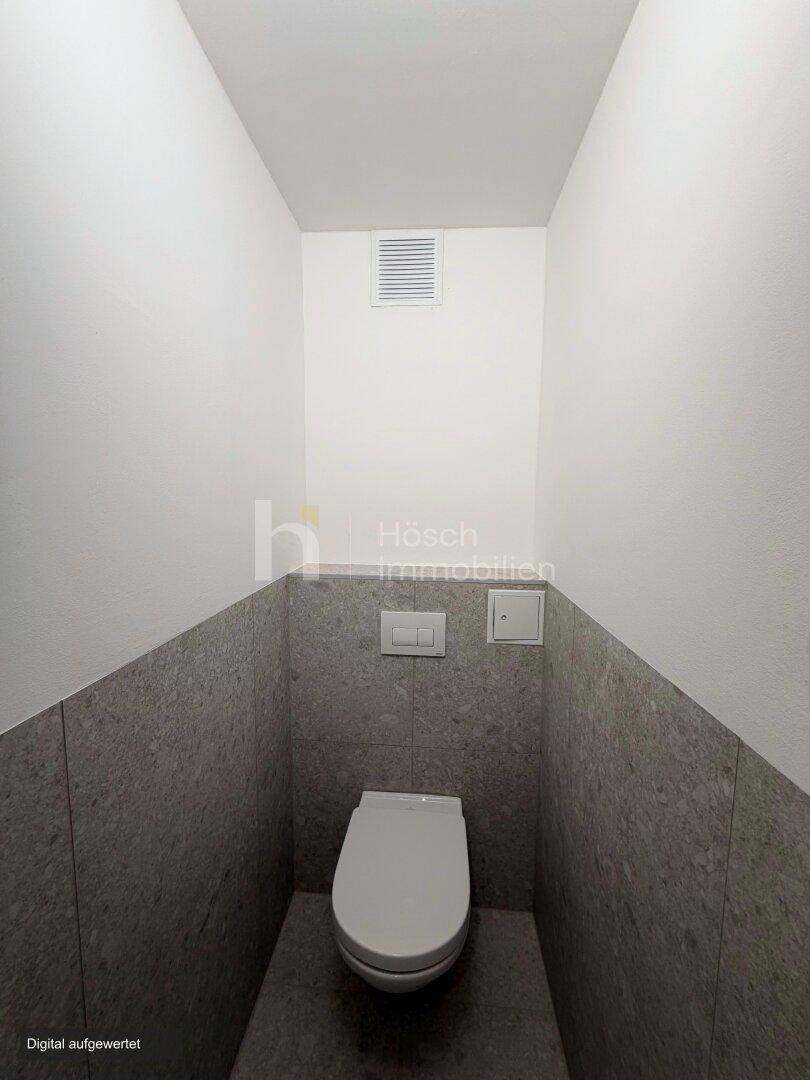 Separates WC mit Wand-WC, grauen Fliesen im unteren Bereich und Lüftungsgitter.