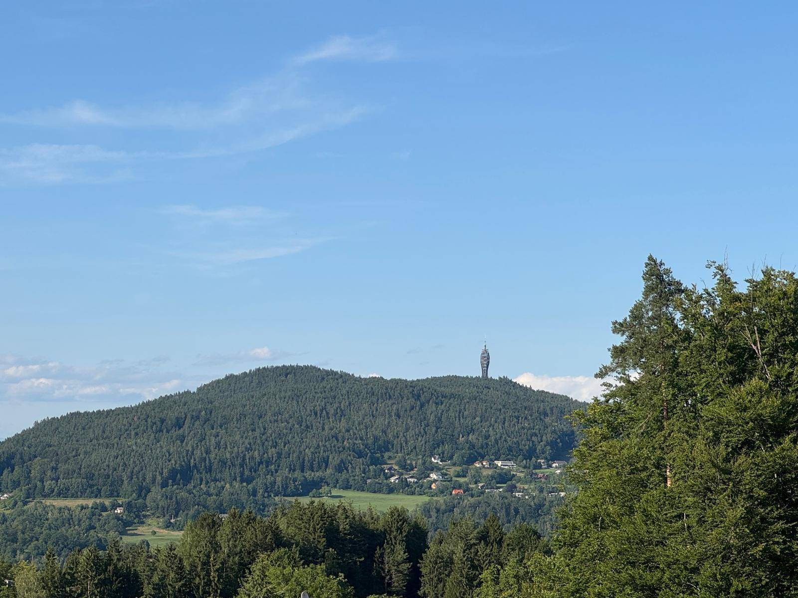 Fernblick auf bewaldete Berge mit einem markanten Sendeturm am Horizont unter blauem Himmel.