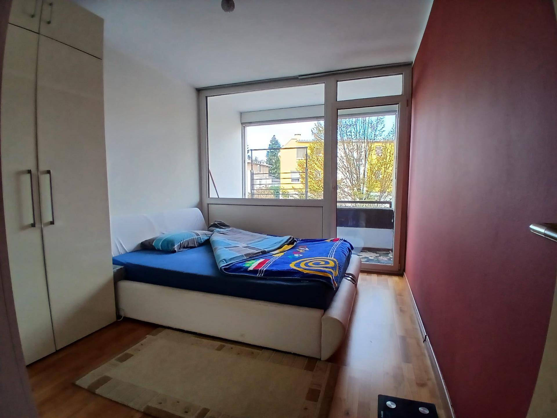 Schlafzimmer mit Holzboden, roter Akzentwand und direktem Zugang zu einem Balkon. Viel Tageslicht durchflutet den Raum.