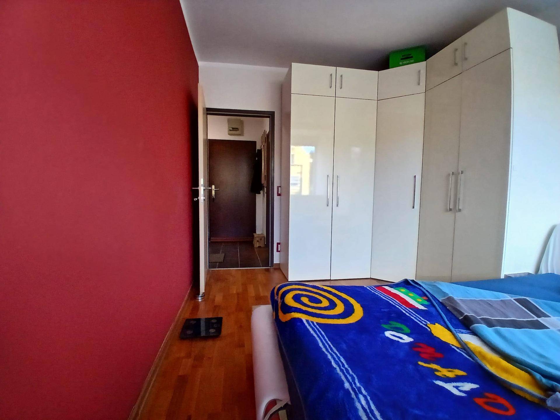 Schlafzimmer mit roter Akzentwand, Holzboden und einem praktischen weißen Eckkleiderschrank. Einladende und helle Gestaltung.