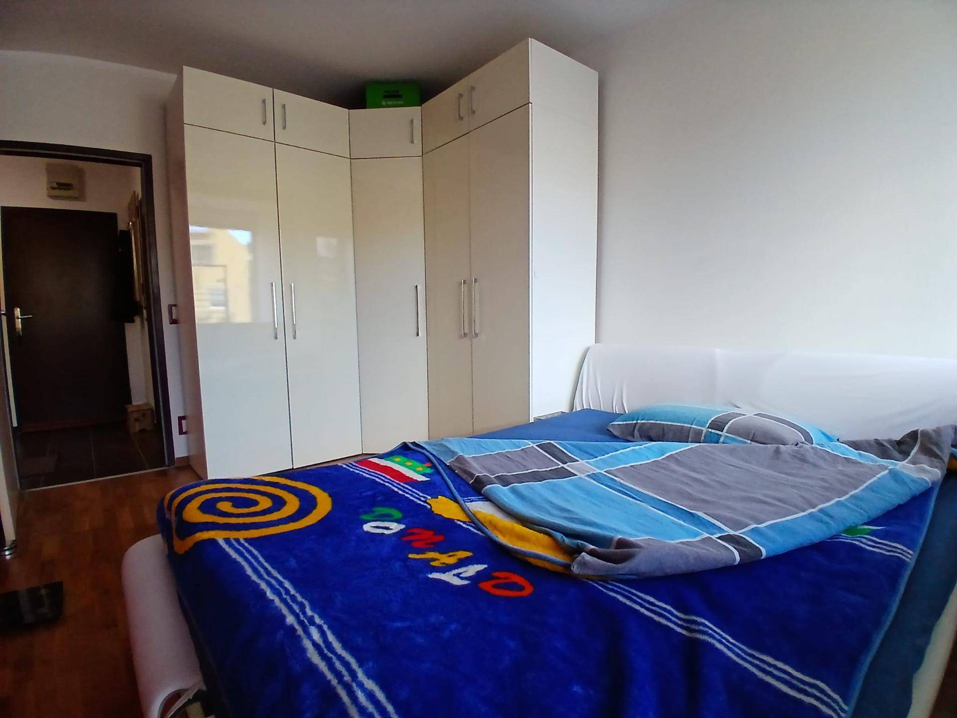 Schlafzimmer mit weißem Eckkleiderschrank, Holzboden und farbiger Bettwäsche. Der Raum wirkt funktional und hell.