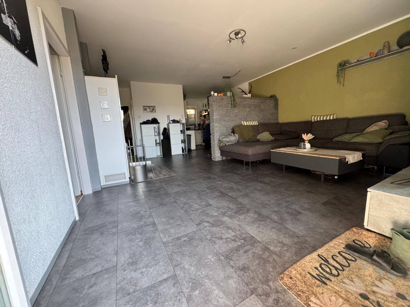 Geräumiges Wohnzimmer mit Fliesenboden, grüner Akzentwand und Blick auf den Eingangsbereich.