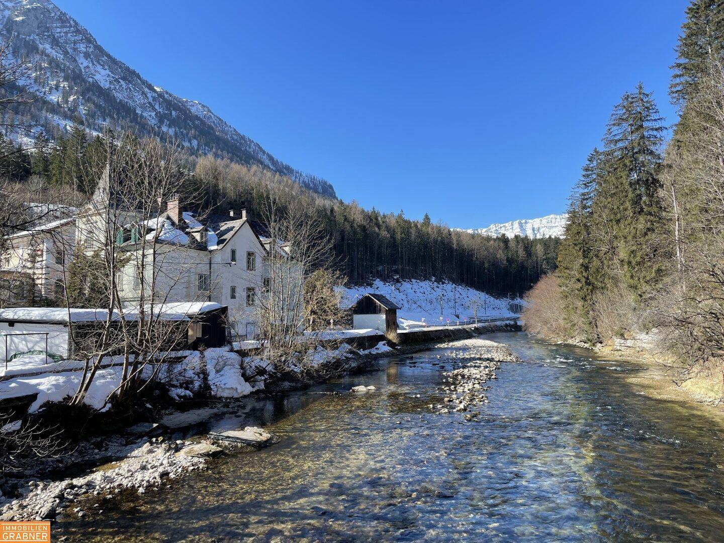 Weißes Gebäude am Flussufer in einer verschneiten Berglandschaft unter klarem Winterhimmel.