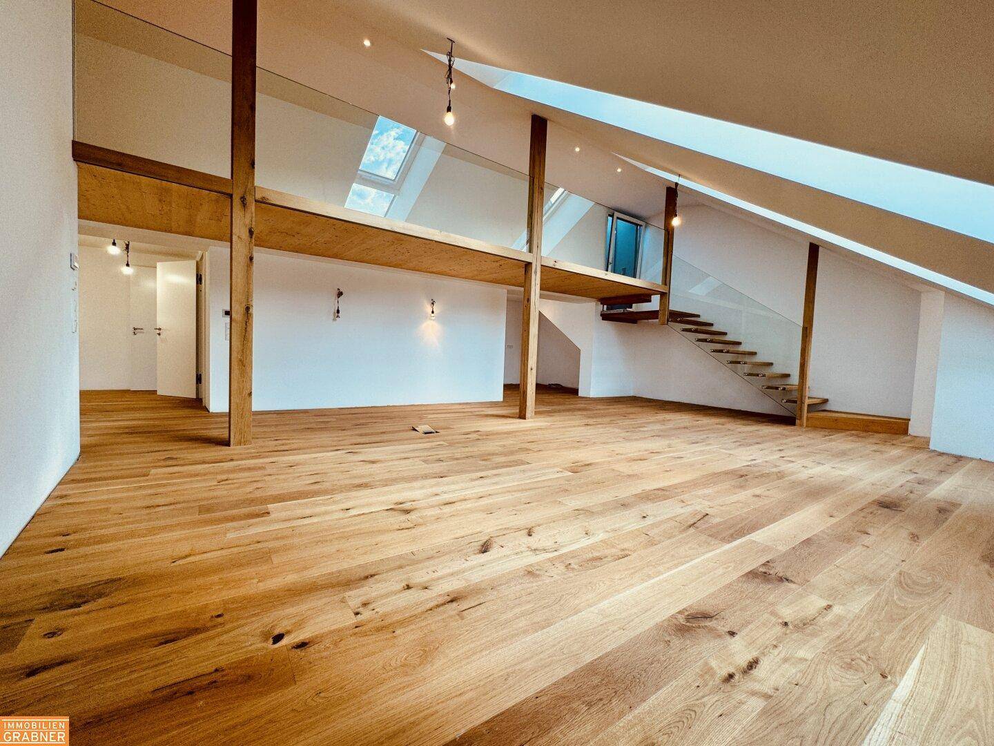 Offener Dachbodenbereich mit Holzboden, Holzbalken und einer eleganten Treppe mit Glasgeländer.