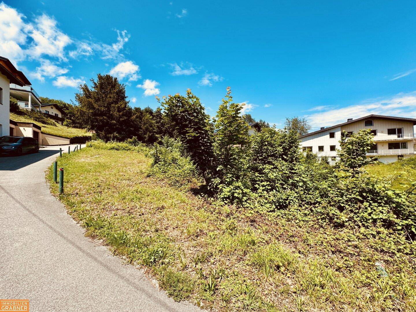 Unbebautes Grundstück mit Gras und Büschen, neben einem asphaltierten Weg und umgeben von Wohnbebauung.