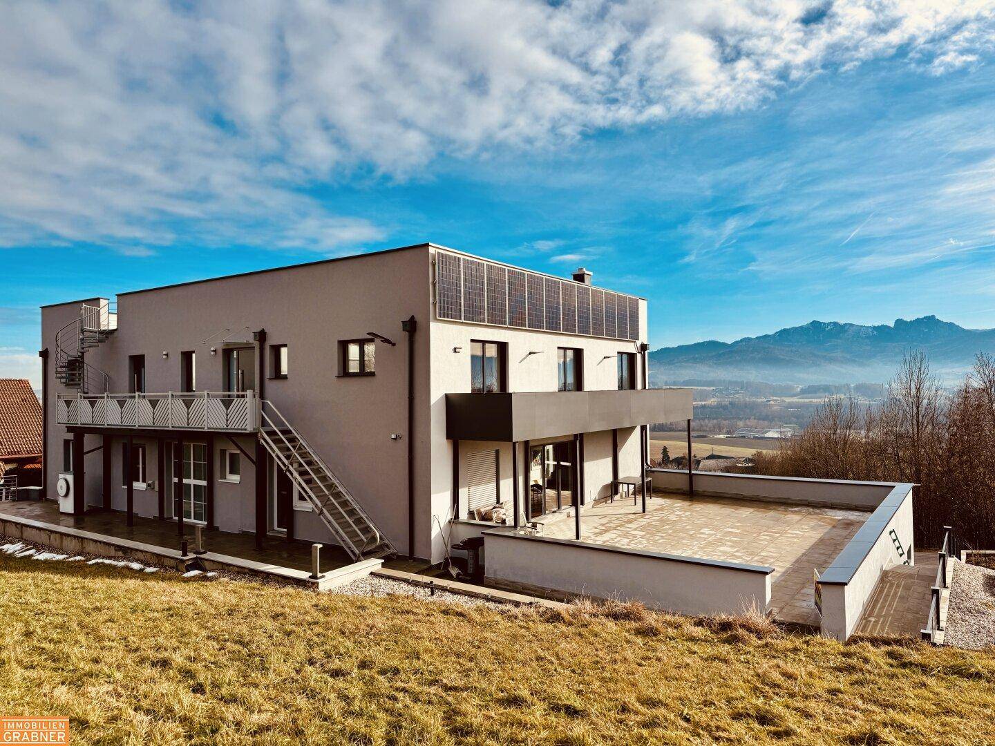 Modernes Gebäude mit großer Terrasse, Photovoltaikanlage auf dem Dach und Bergblick.