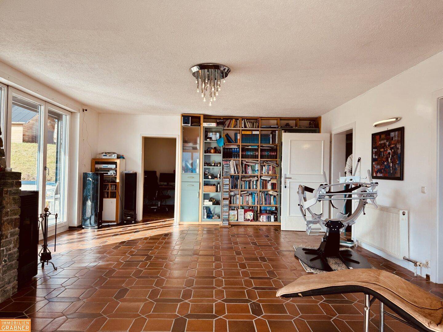 Heller Wohnbereich mit Terrakotta-Fliesenboden, Bücherregal und Zugang zu einem weiteren Raum.