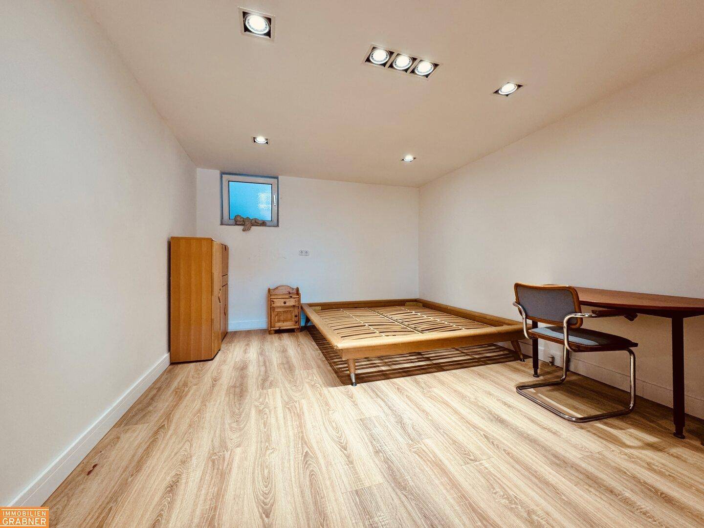 Leeres Schlafzimmer mit Laminatboden, kleinem Fenster und Einbaustrahlern.