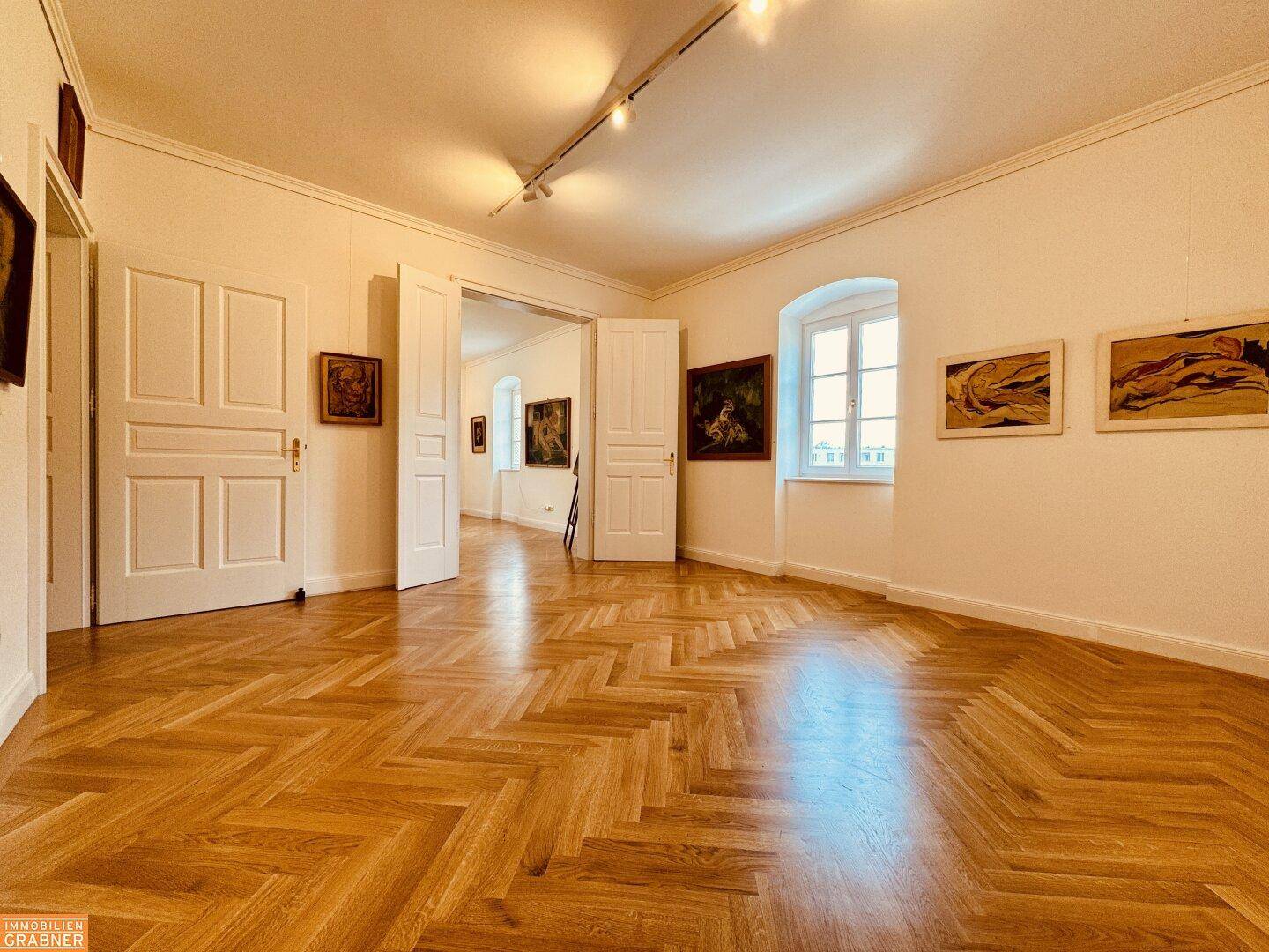 Geräumiger Raum mit Fischgrätparkett, hohen Türen und Kunstwerken an den Wänden.