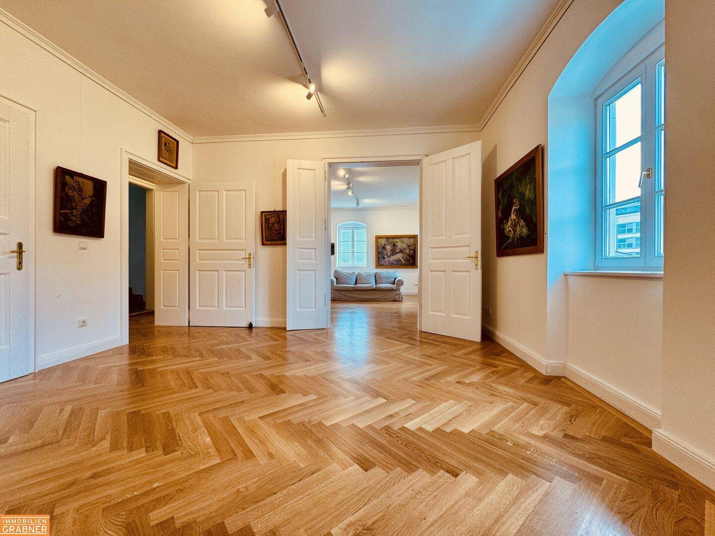 Langer Flur mit Fischgrätparkett und hohen Türen, der einen Blick in den nächsten Raum bietet.