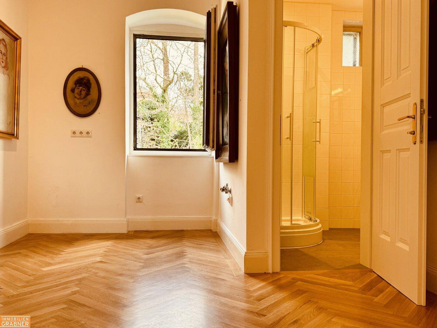 Wohnraum mit Fischgrätparkett und Blick in ein Badezimmer mit Duschkabine.