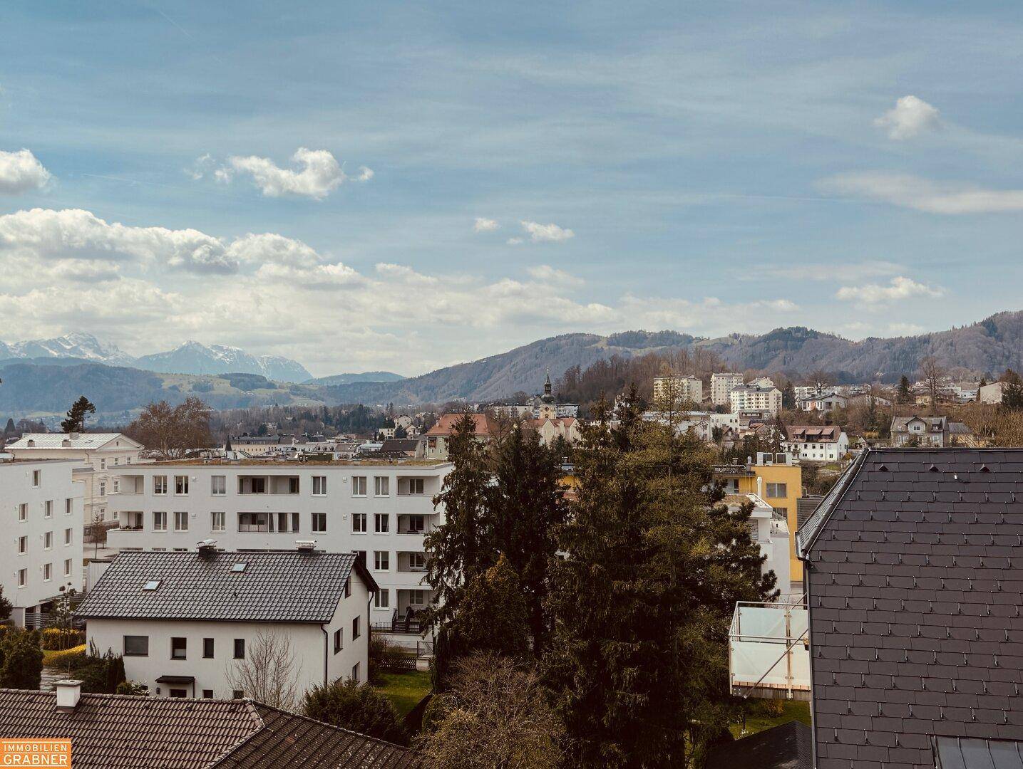 Panoramablick über eine Stadtlandschaft mit Bergen im Hintergrund unter bewölktem Himmel.