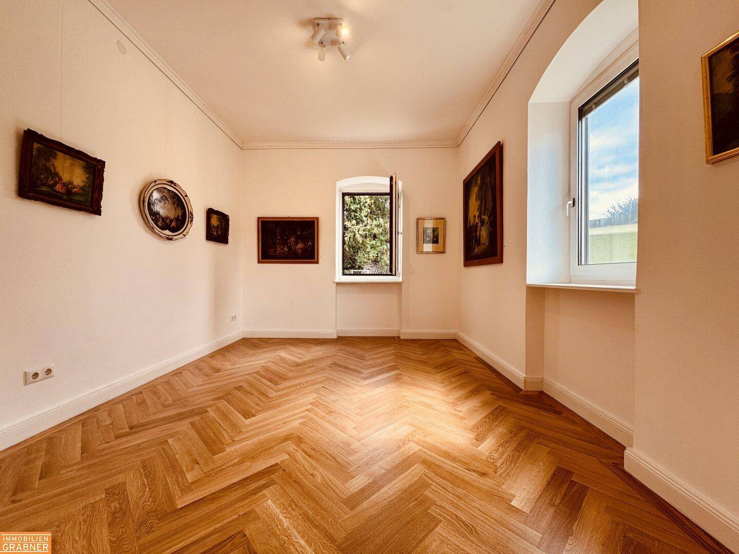 Heller Raum mit Fischgrätparkett, hohen Decken und mehreren Fenstern, dekoriert mit Kunstwerken.