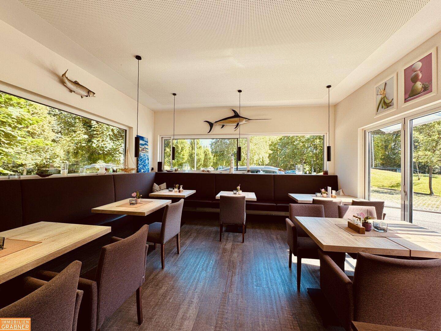 Heller Restaurant-Innenraum mit Holzböden, bequemen Sitzbänken und großen Fenstern mit Blick ins Grüne.