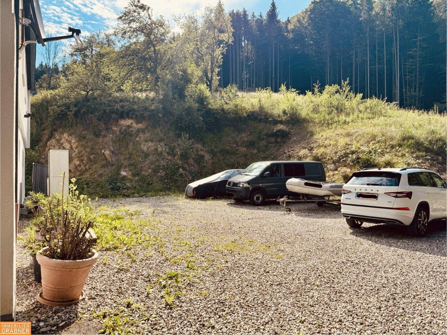 Kiesparkplatz mit mehreren Fahrzeugen und Blick auf bewaldete Hügel.