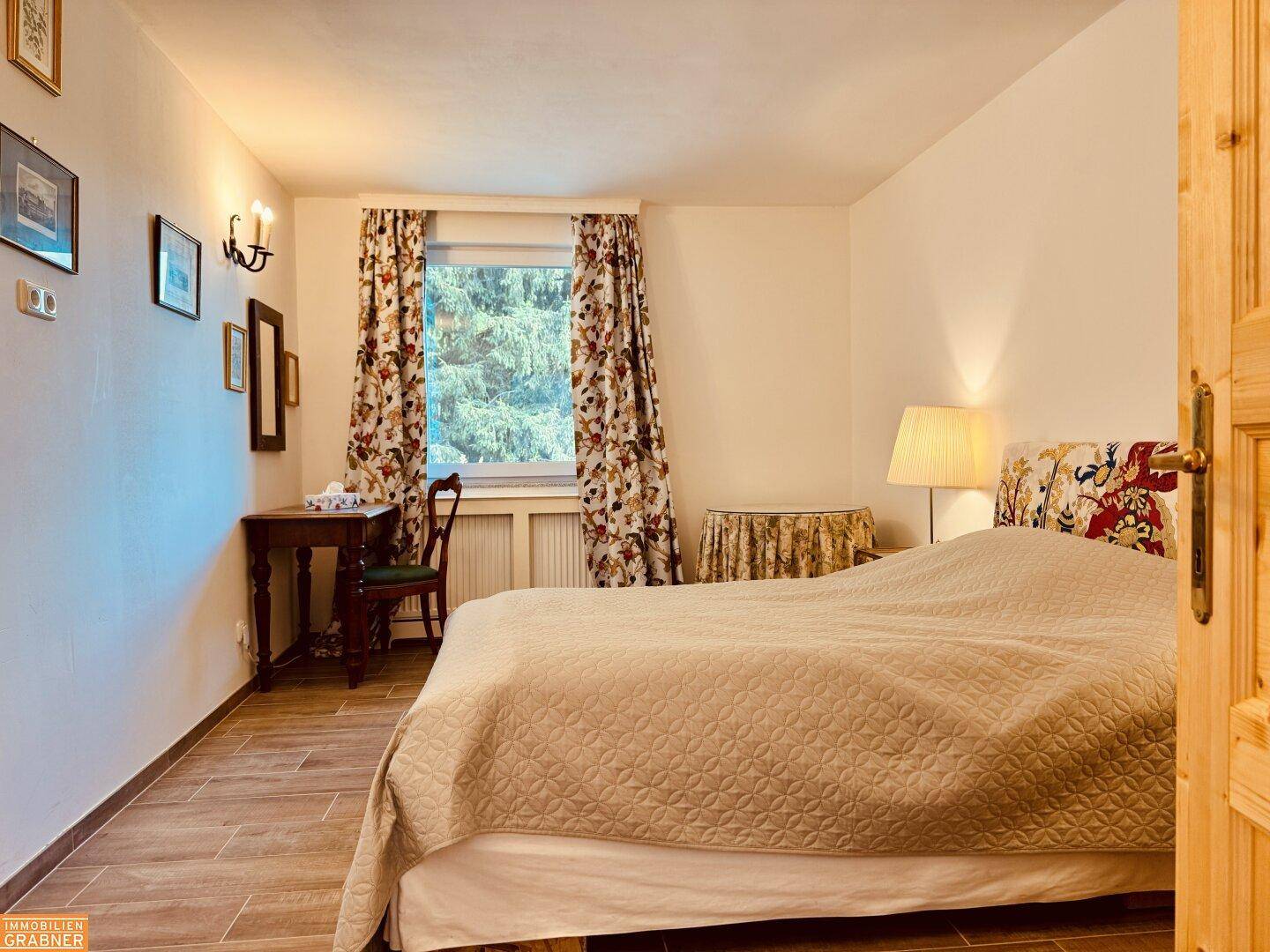 Gemütliches Schlafzimmer mit Holzboden, floralen Vorhängen und traditionellen Möbeln.