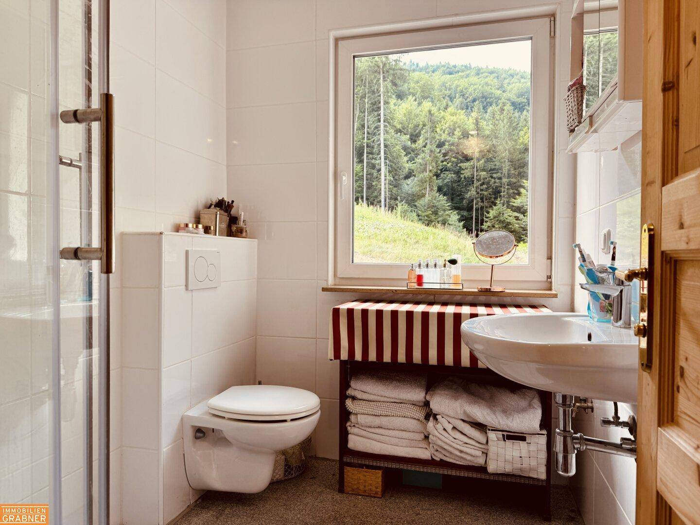 Helles Badezimmer mit WC, Waschbecken und Duschkabine, mit Fenster und Blick ins Grüne.