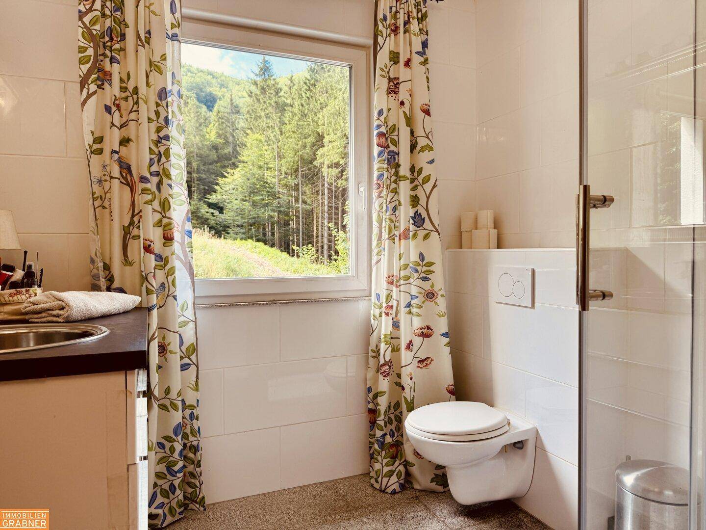 Badezimmer mit WC, Waschbecken und Duschkabine, mit floralen Vorhängen und Fensterblick.