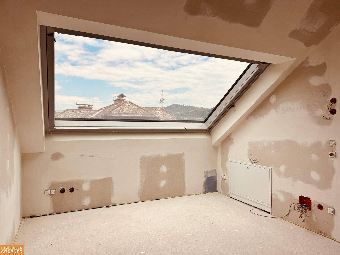 Unfertiger Innenraum mit großem Dachfenster, das einen weiten Blick auf Dächer und Hügel bietet.