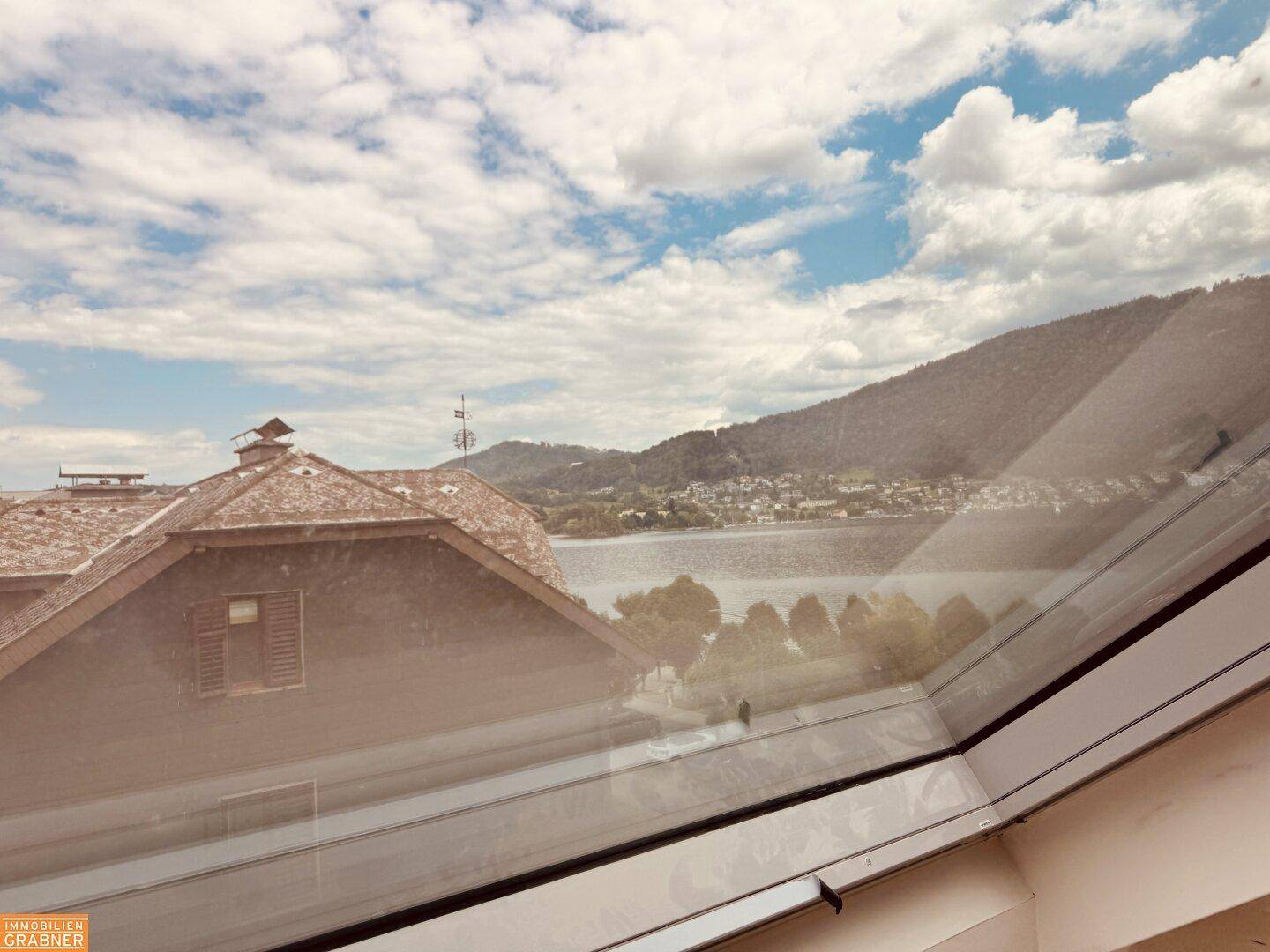 Panoramablick aus dem Dachfenster auf einen malerischen See und die umliegenden grünen Berge.