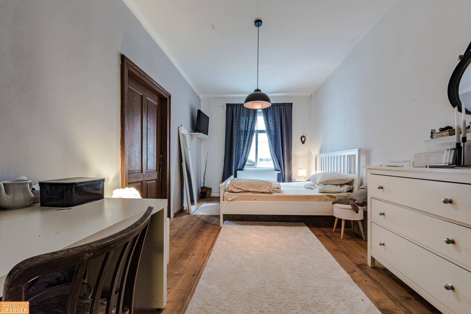 Schlafzimmer mit Holzboden, weißem Bett und dunklen Vorhängen, bietet eine ruhige Rückzugsmöglichkeit.