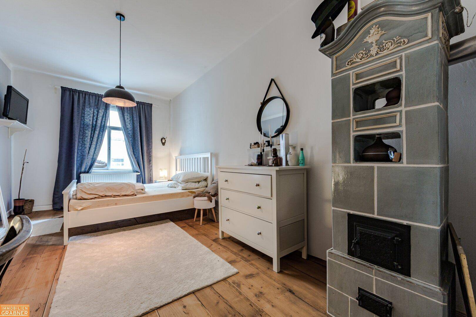 Schlafzimmer mit Holzboden, weißem Bett und einem traditionellen Kachelofen, schafft eine warme Atmosphäre.
