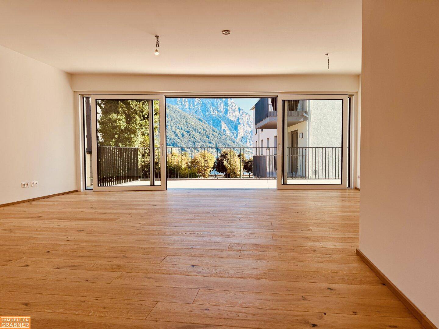 Helles, leeres Wohnzimmer mit Eichenparkett und großen Schiebetüren zum Balkon mit Bergblick.