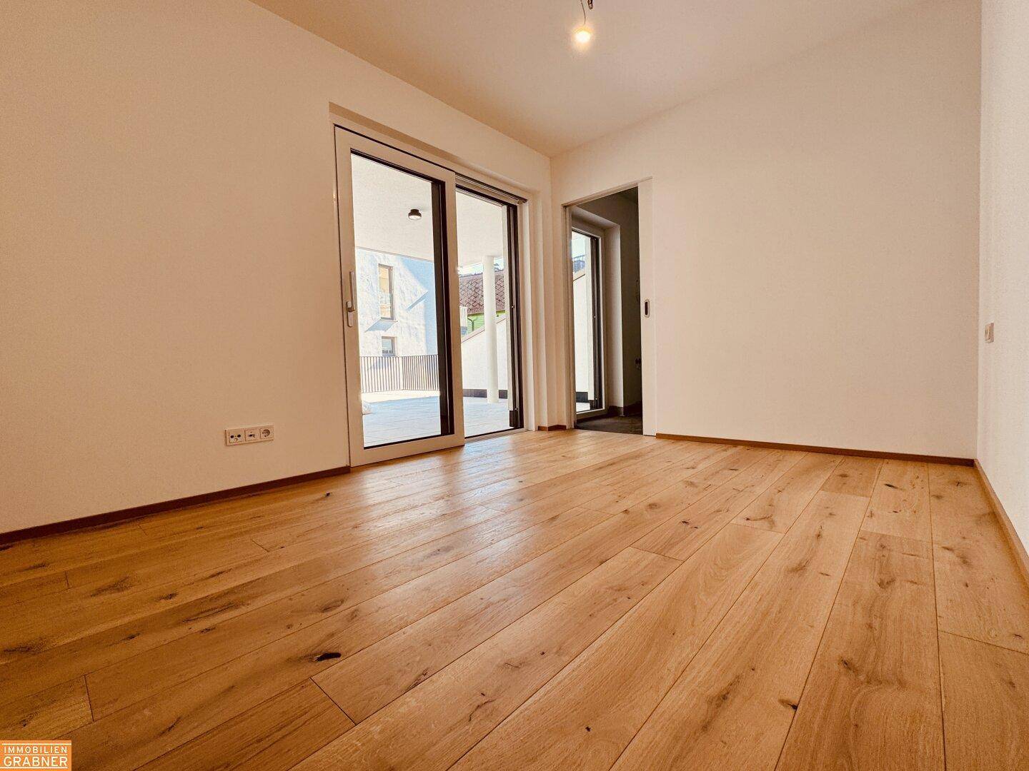 Leeres Zimmer mit hellem Holzboden und Zugang zu einem Außenbereich durch eine große Glastür.