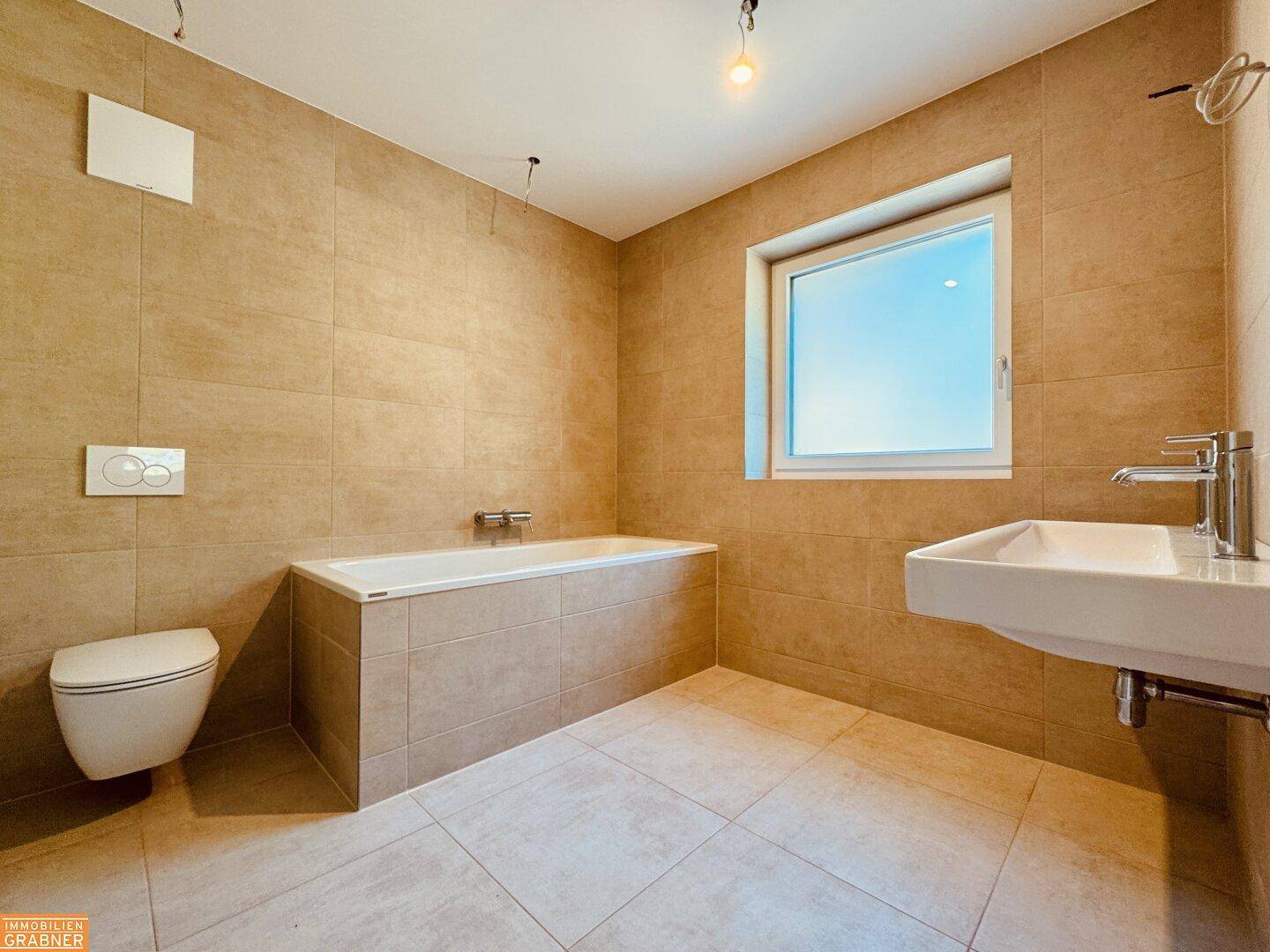 Modernes Badezimmer mit beigefarbenen Fliesen, Badewanne, wandhängendem WC und Fenster.