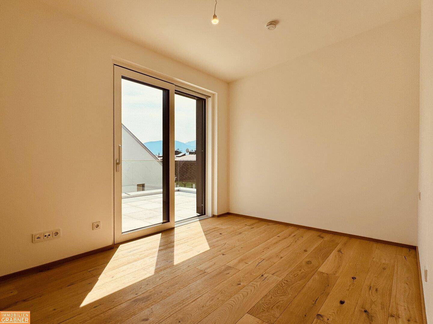 Helles Zimmer mit Holzboden und Zugang zu einem kleinen Balkon oder einer Terrasse mit Weitblick.