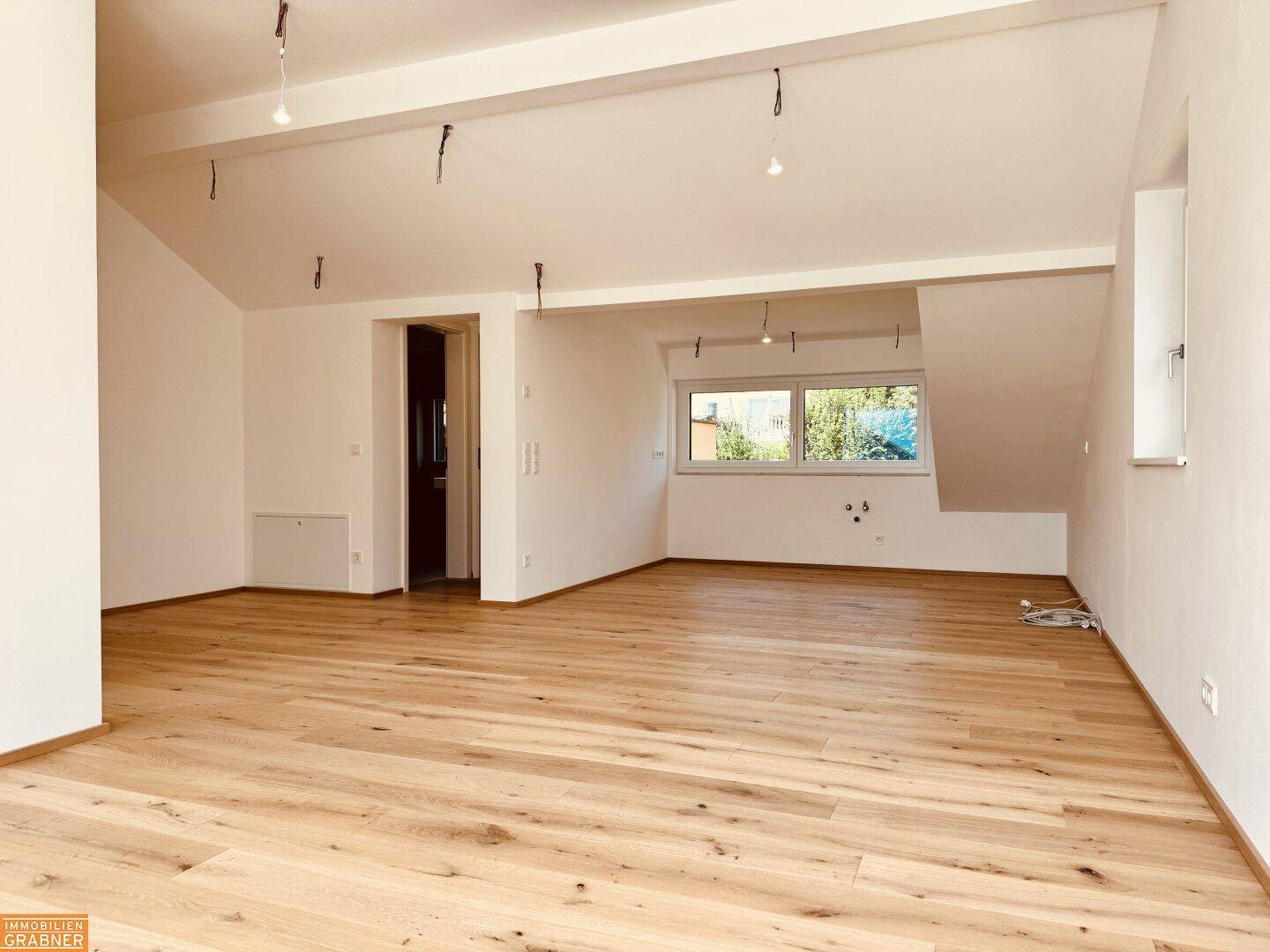 Geräumiger Raum im Dachgeschoss mit Holzboden, schrägen Decken und Fenster mit Blick ins Grüne.