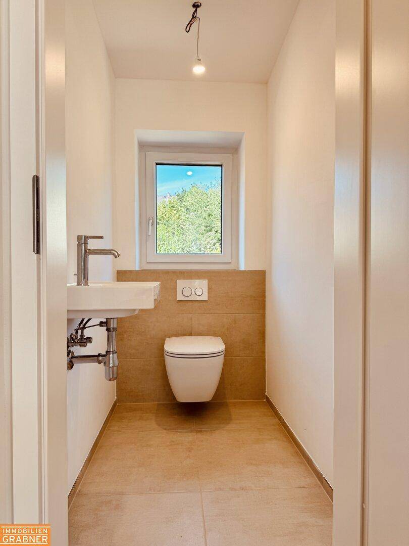 Gäste-WC mit beigefarbenen Fliesen, wandhängendem WC, Waschbecken und Fenster für Tageslicht.