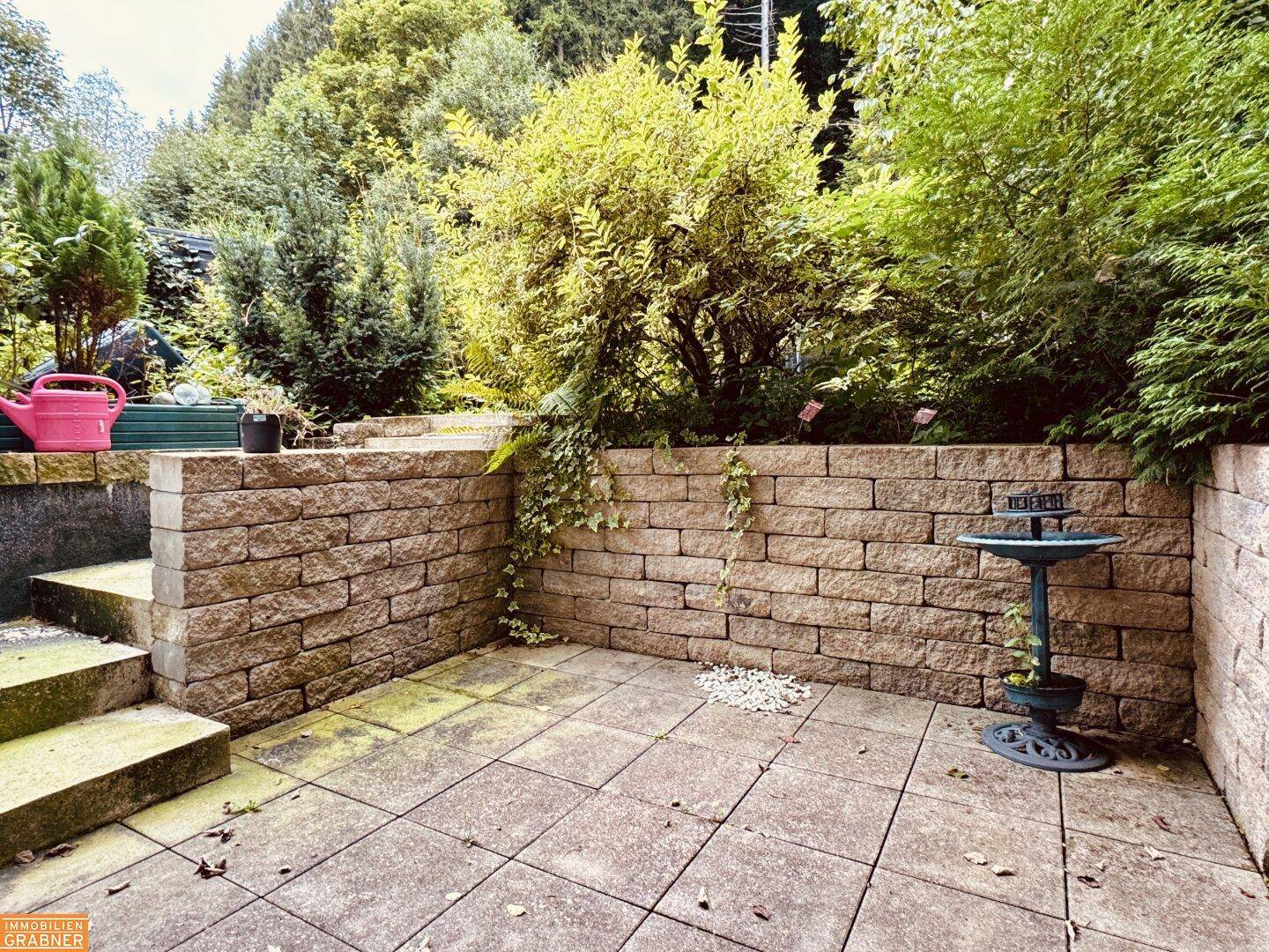 Steinplatten-Terrasse mit Natursteinmauer und üppiger Bepflanzung im Hintergrund.
