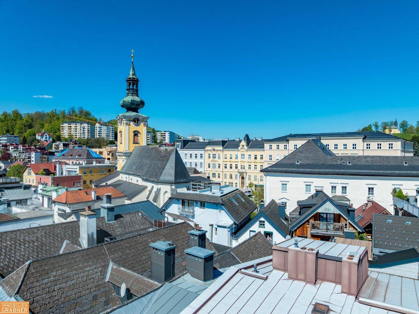 Weitläufiger Blick über die Dächer der Stadt mit historischem Kirchturm und grünen Hügeln.