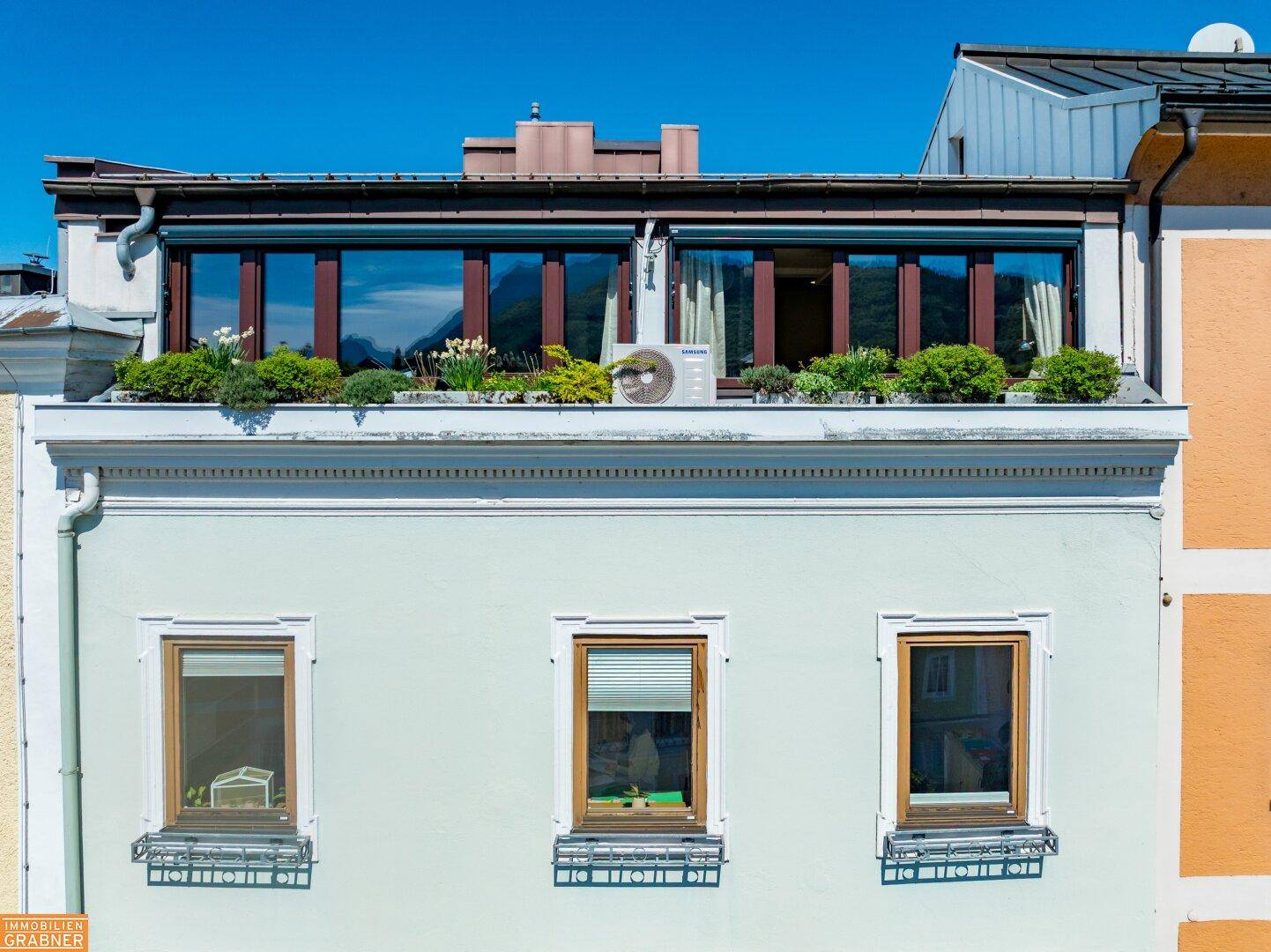 Obere Etage des Gebäudes mit großen Fenstern und einer Dachterrasse mit Bepflanzung.