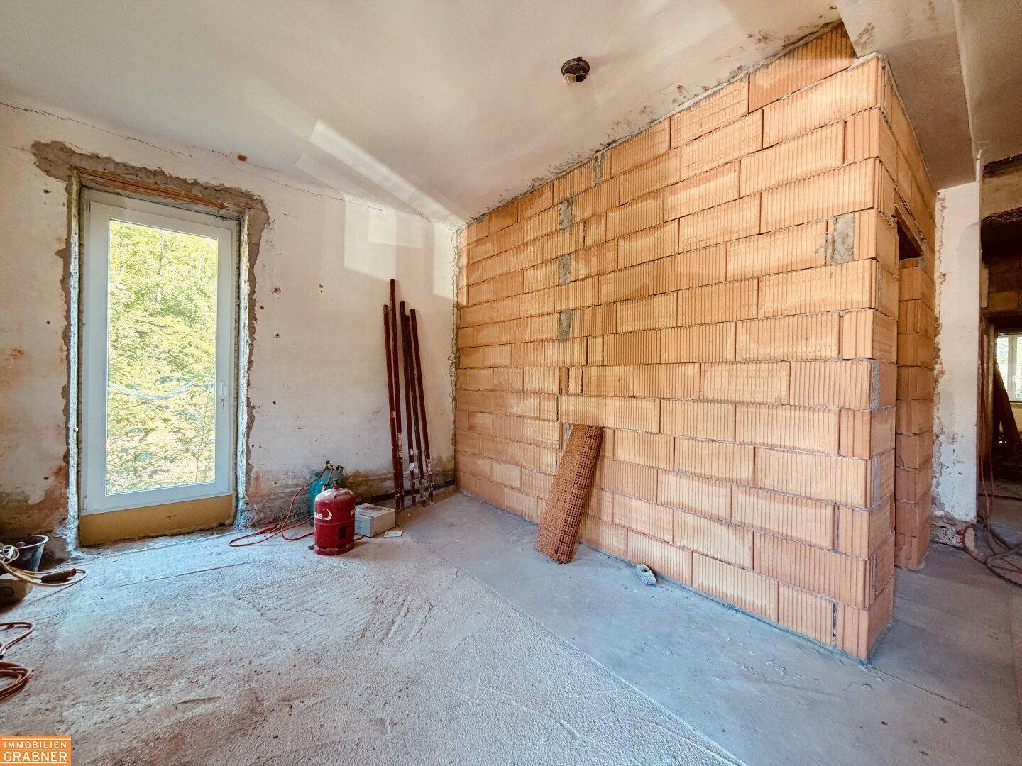 Unfertiger Innenraum mit Ziegelwänden und einem großen Fenster, bereit für den Ausbau.