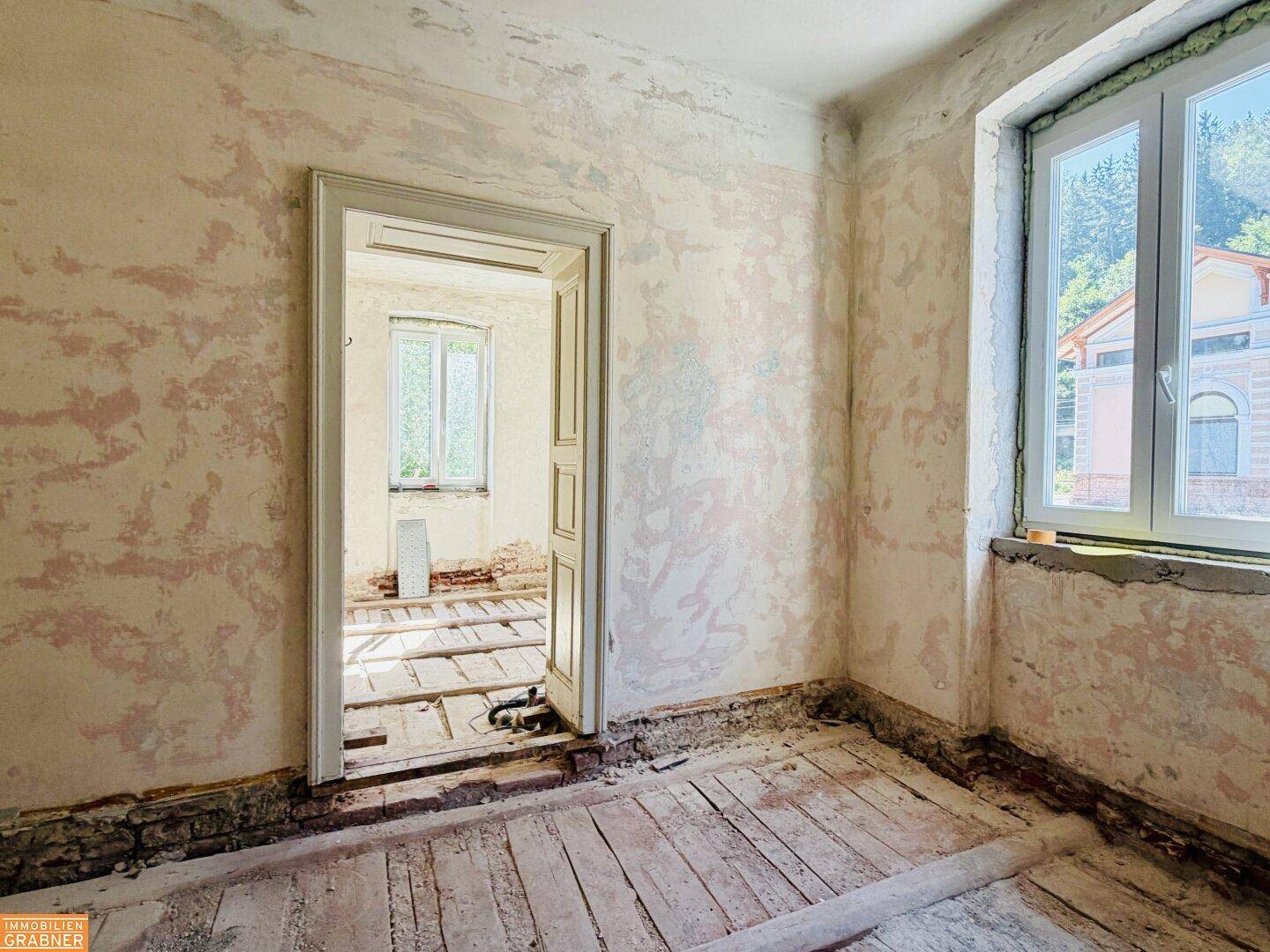 Zimmer im Rohzustand mit freigelegten Holzdielen und Blick ins Grüne durch das Fenster.