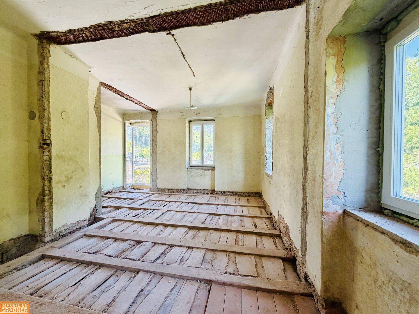 Leerer Raum in Renovierungszustand mit freiliegenden Holzdielen und abblätternden Wänden.