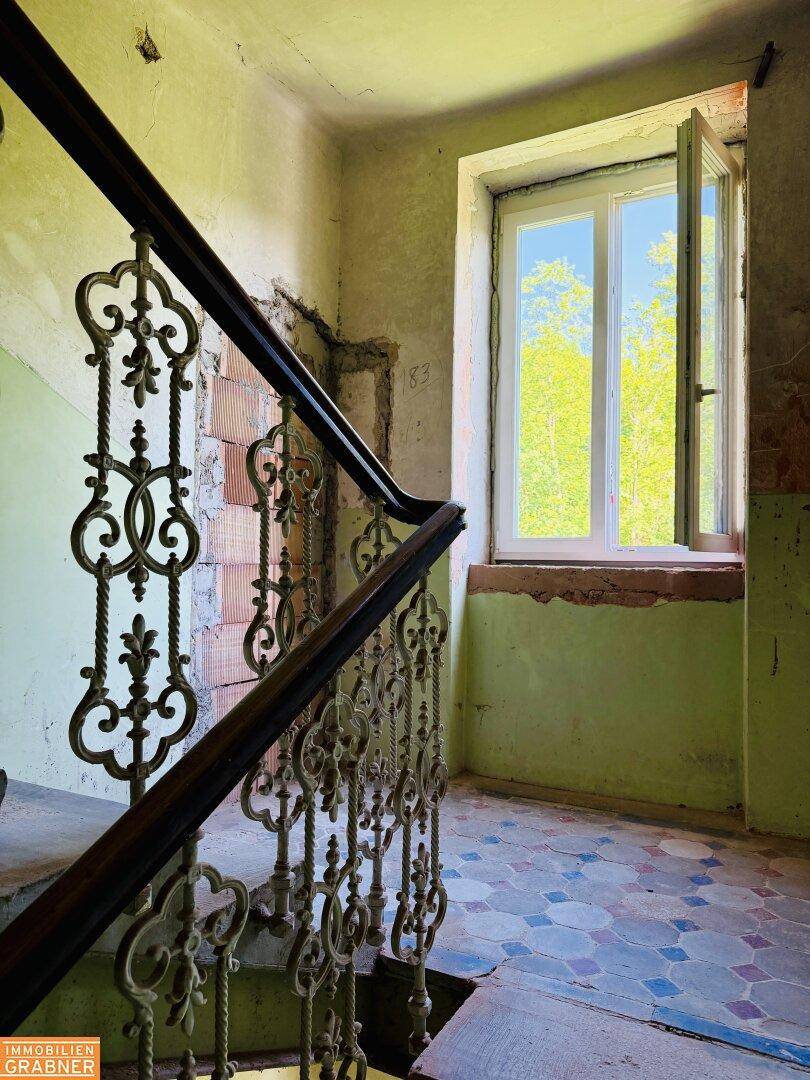 Altes Treppenhaus mit schmiedeeisernem Geländer, gemusterten Fliesen und einem Fenster mit Blick ins Grüne.